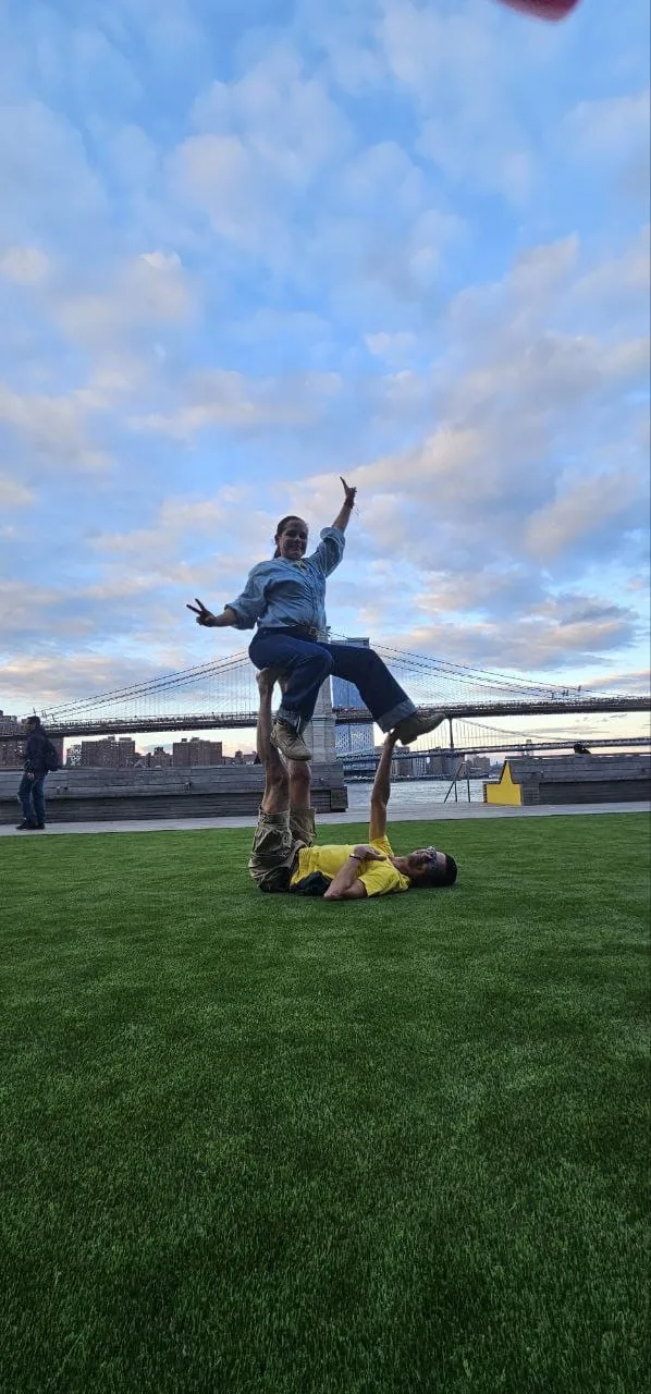 Ernesto Hoyos sosteniendo con los pies a una alumna en una postura de acroyoga, con el puente de Brooklyn al fondo